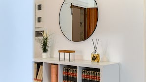 A white console table with a mirror above it, a potted plant, a small wooden stool, and a bottle of reed diffuser. Below the table, there are shelves with books and decorative items. - Monteverde Marí House Vacation Home with Wi-Fi (Rome)