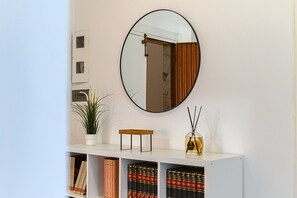 Ein weißer Konsolentisch mit einem Spiegel darüber, einer Topfpflanze, einem kleinen Holzschemel und einer Flasche mit Schilferdiffusor. Darunter befinden sich Regale mit Büchern und dekorativen Gegenständen.