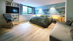 Une chambre à coucher avec un lit, un fauteuil à bascule, une bibliothèque, un téléviseur et une grande fenêtre.