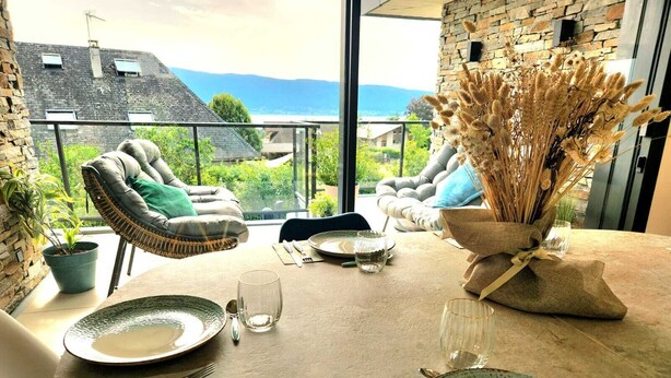 Un coin repas avec une table dressée pour deux personnes, une chaise en osier, une plante en pot et une vue sur l’extérieur.