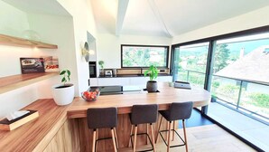 Une cuisine moderne avec un îlot central en bois, des tabourets de bar, une grande fenêtre et une vue sur l’extérieur.