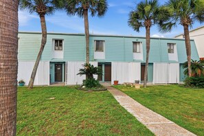 A two-story building with a light blue exterior, a front entrance, and a small garden area with palm trees and a pathway. - Beachfront Home Steps to Sand Fall Open Oct17 (Panama City Beach)