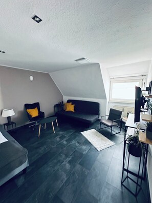 A modern loft-style room with a dark grey sofa, a small black chair, a white desk, and a large window.