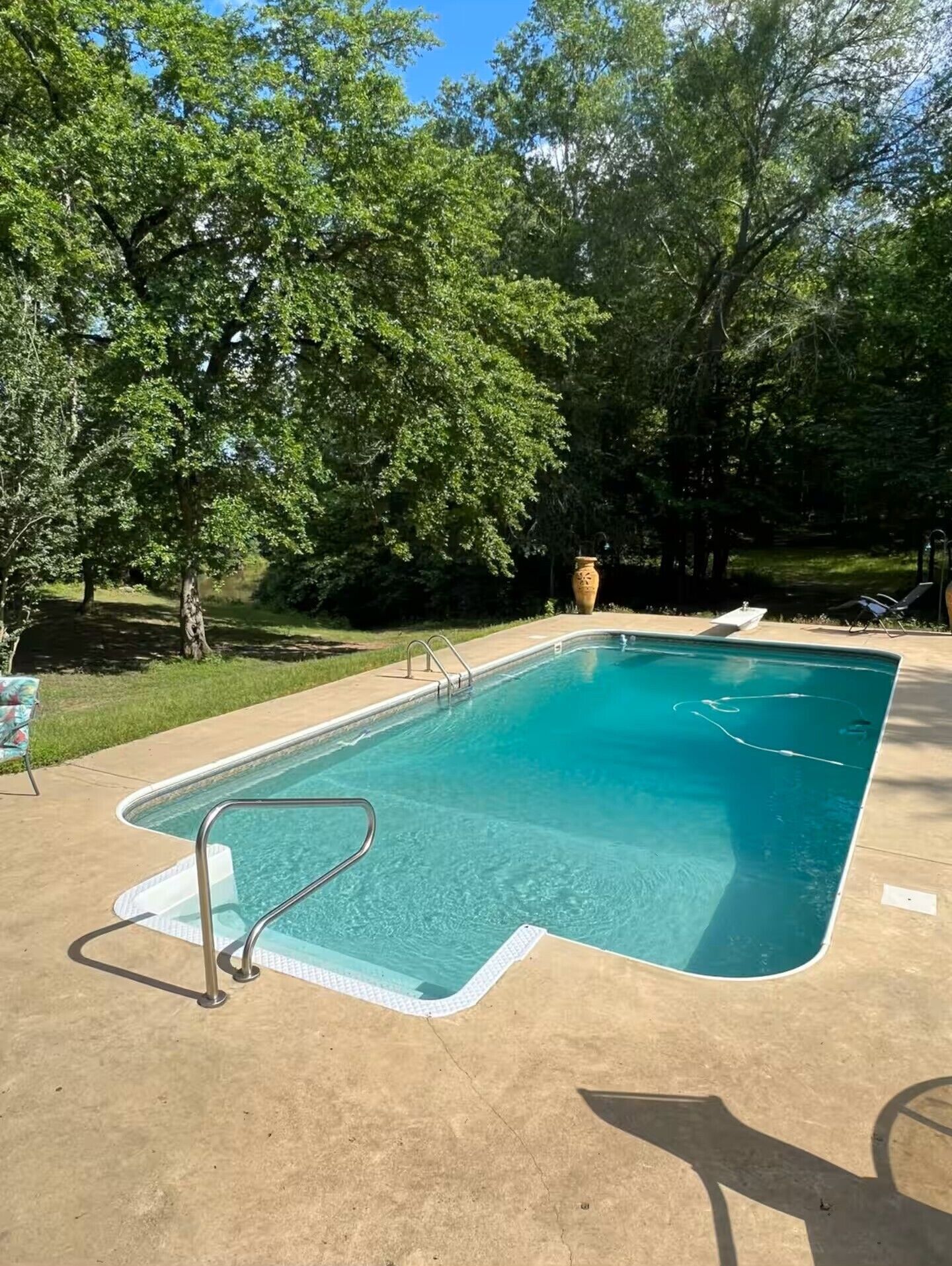 Uma piscina retangular com uma escada de metal, rodeada por árvores e uma área gramada.