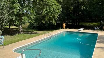 Uma piscina retangular com uma escada de metal, rodeada por árvores e uma área gramada.