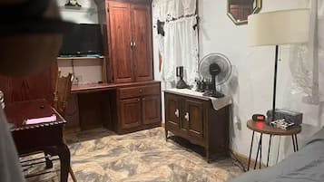 Quarto com guarda-roupa de madeira, uma mesinha com abajur, um ventilador e um relógio em uma mesinha lateral.
