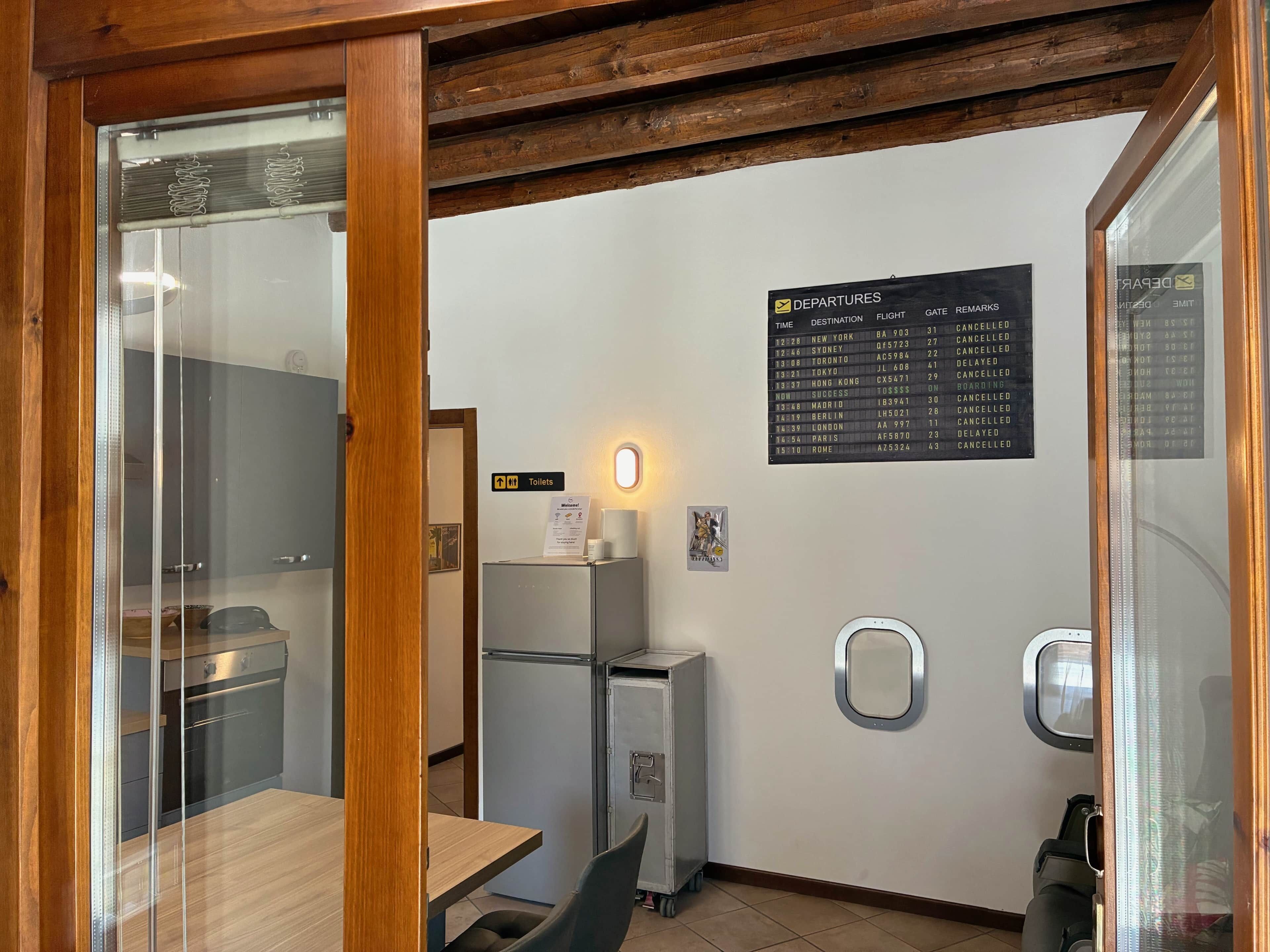 Habitació d'hotel amb sostre de fusta, porta de vidre, taula petita, nevera, armari, senyalitzador de lavabos i panell d'informació de sortides.