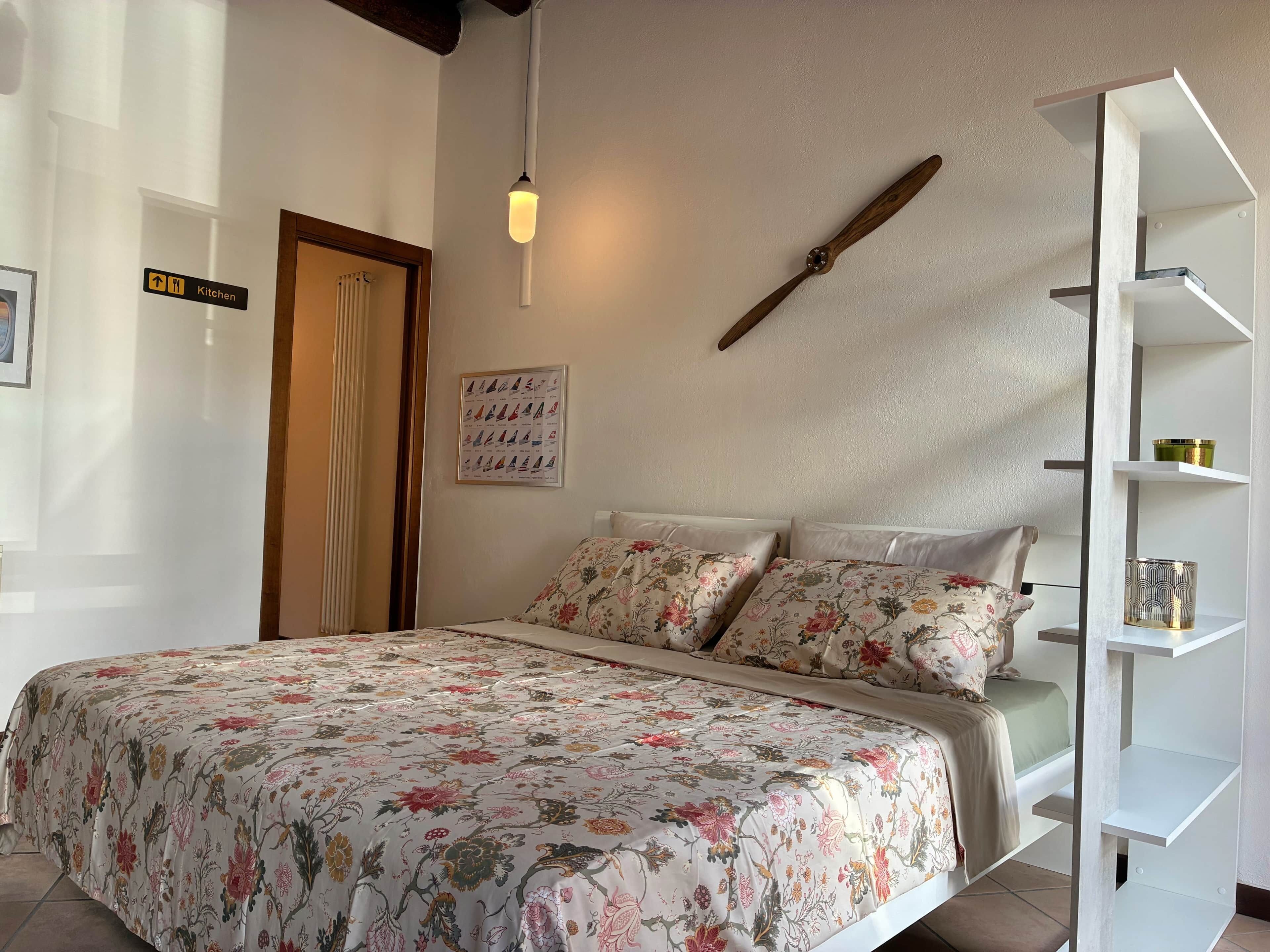 A bedroom with a bed, a nightstand, a wall-mounted shelf, a ceiling fan, and a door.