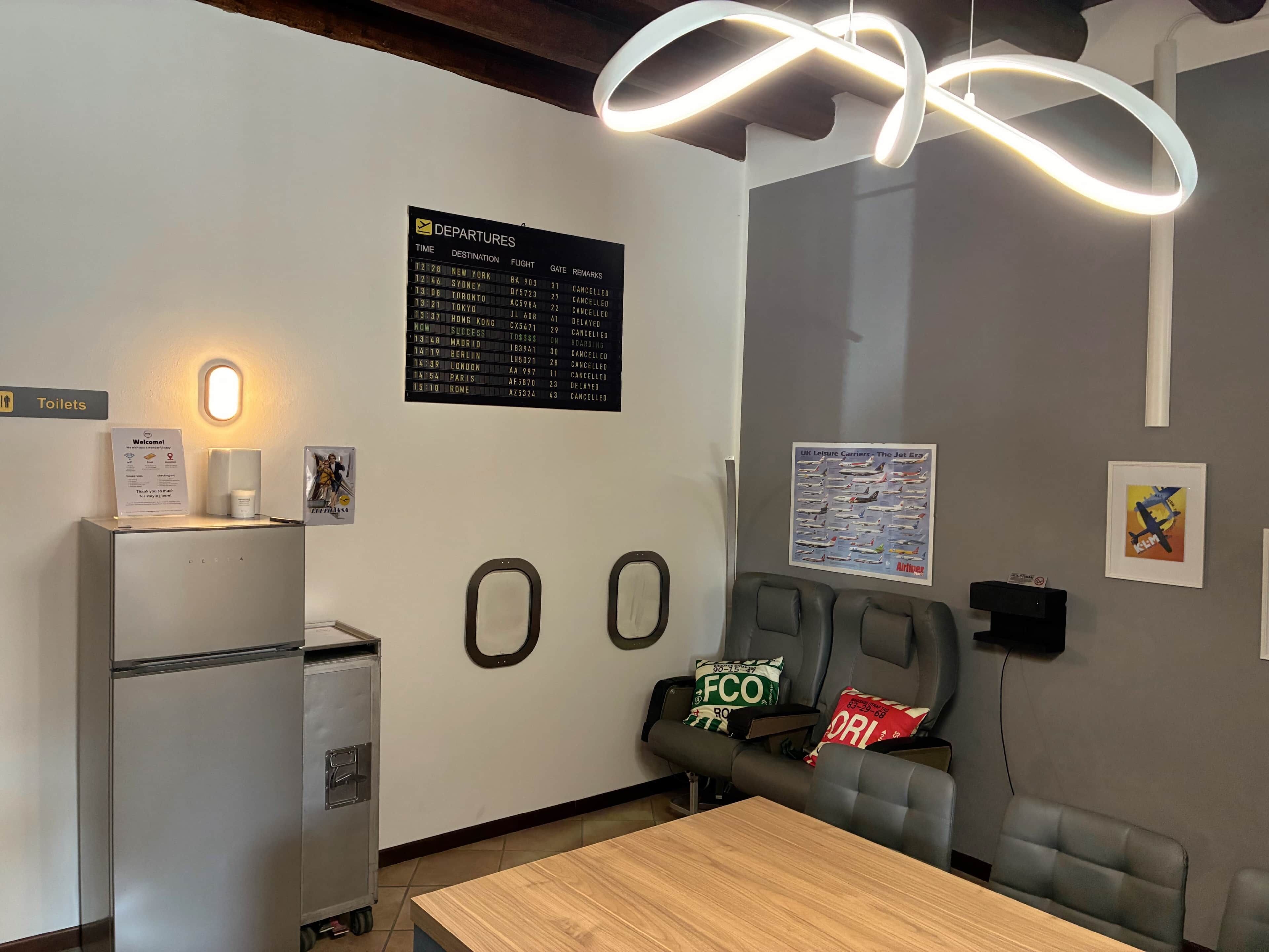 A hotel room with a departure board, a refrigerator, a table, a sofa, and a wall-mounted TV.