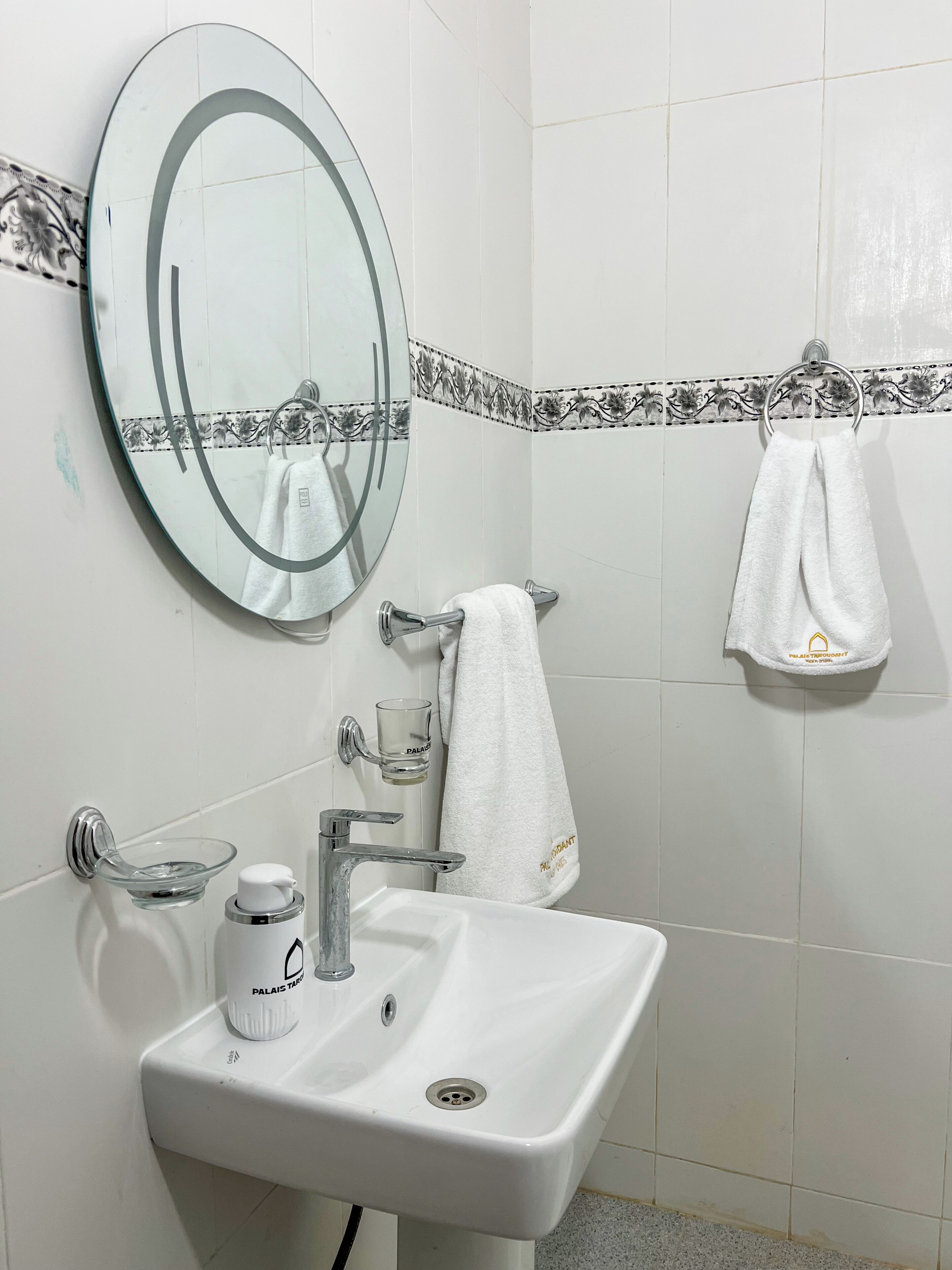 Une salle de bain avec un miroir rond, un lavabo blanc, un distributeur de savon en verre et un porte-serviettes avec deux serviettes.