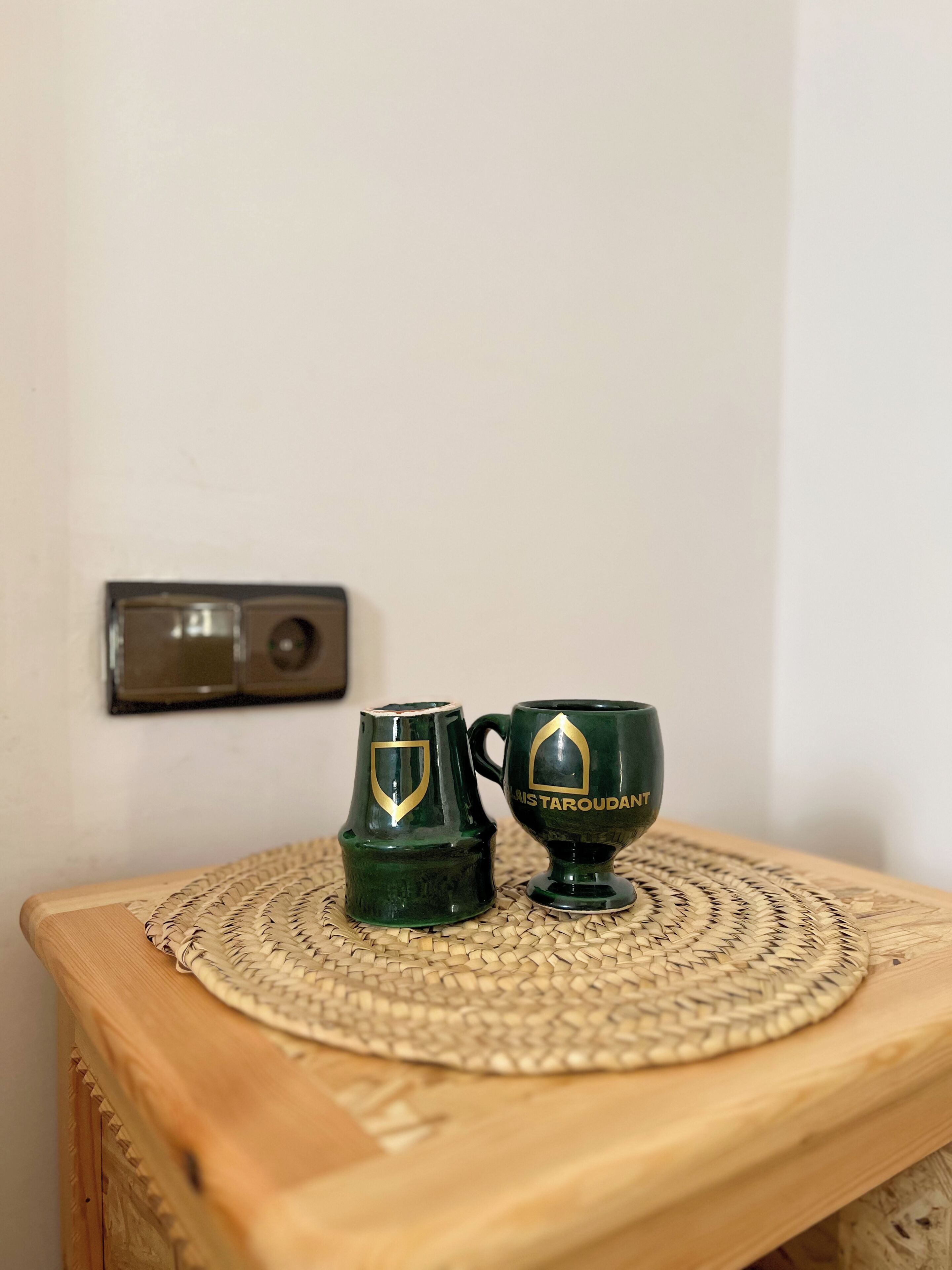 Two green mugs with gold emblems on a woven placemat on a wooden table.