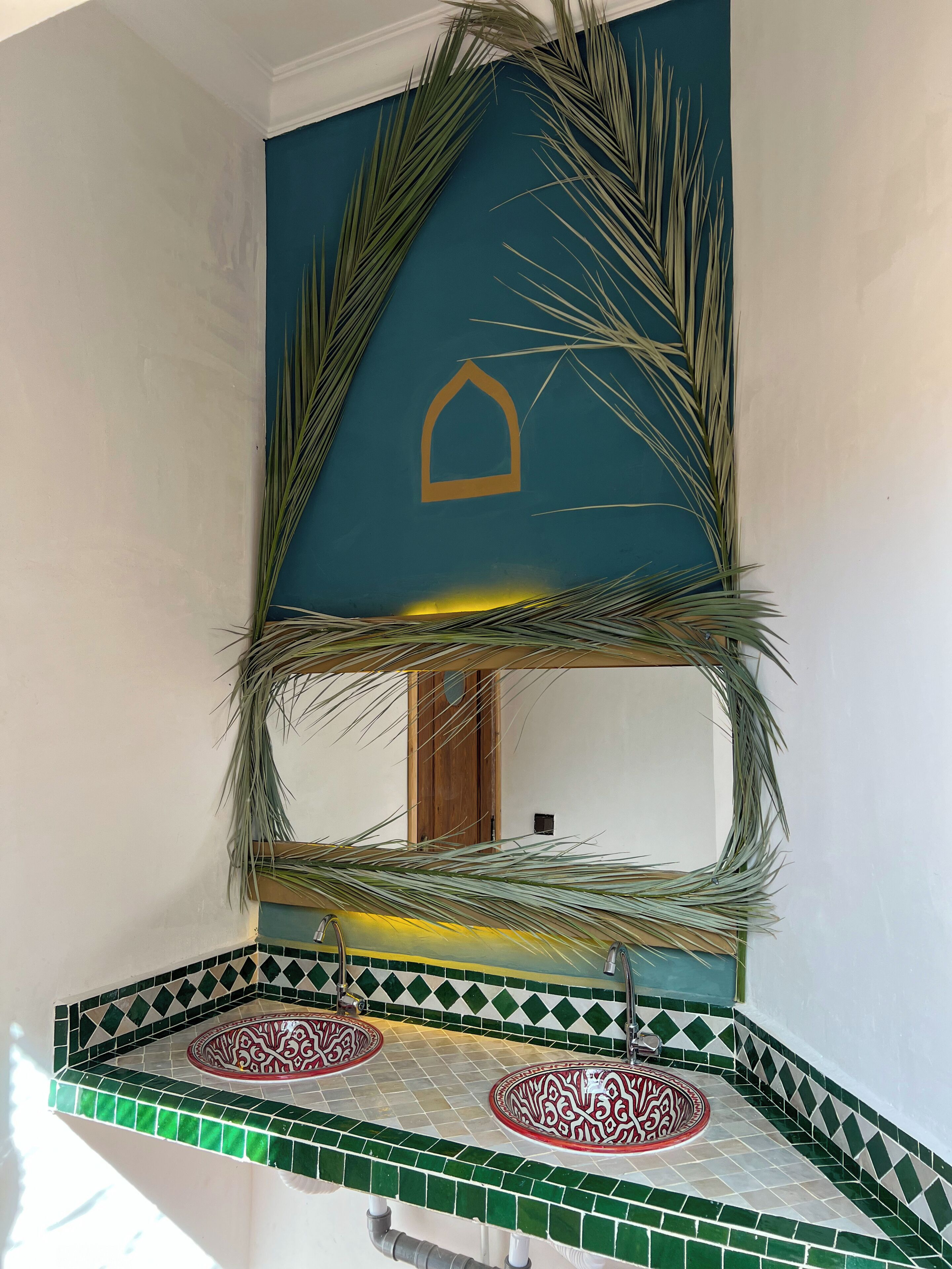 Un baño con zona de lavamanos con azulejos verdes y blancos, un espejo con marco decorativo de hojas de palma y una puerta de madera al fondo.