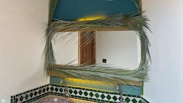 Un baño con zona de lavamanos con azulejos verdes y blancos, un espejo con marco decorativo de hojas de palma y una puerta de madera al fondo.