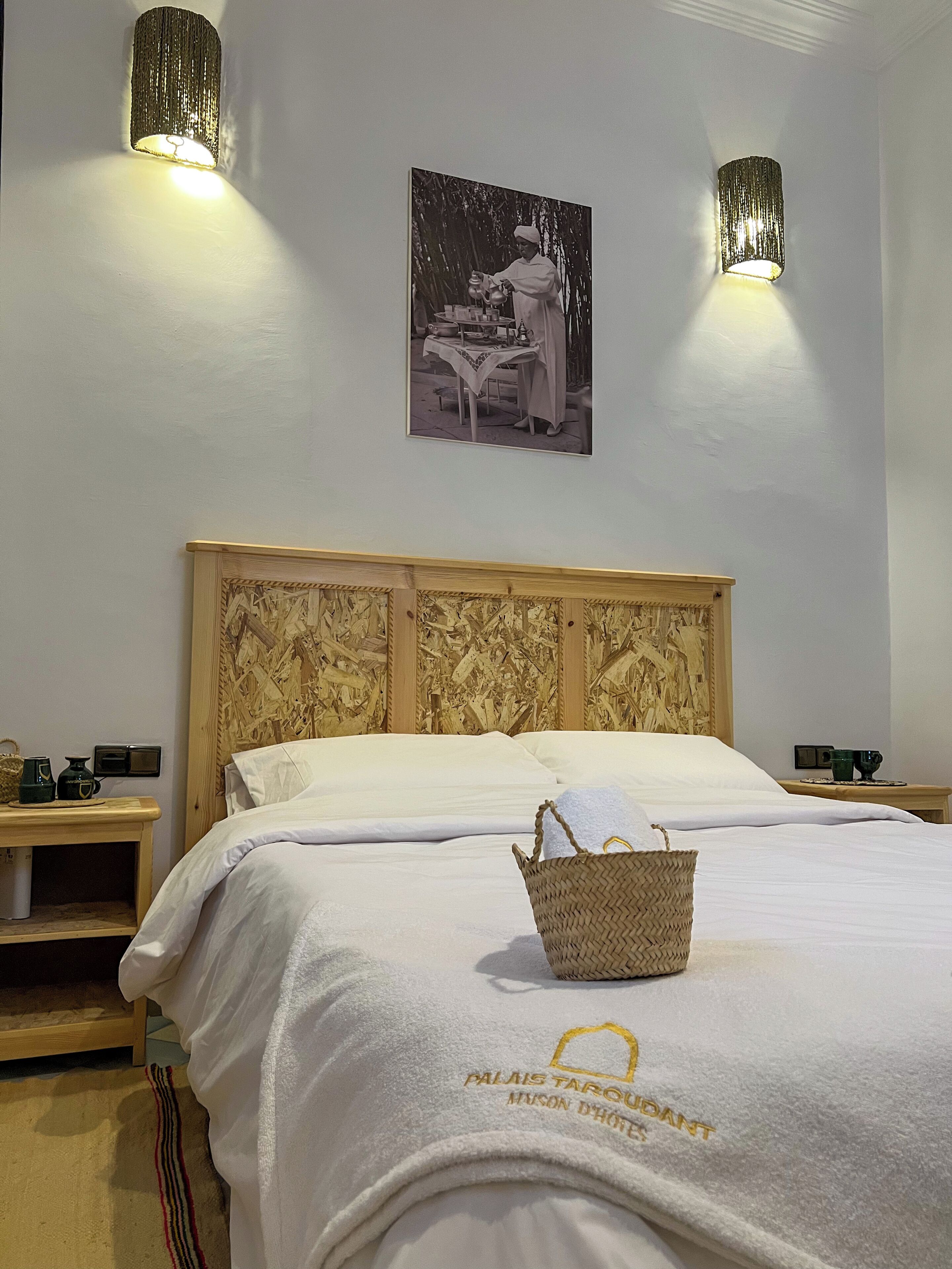 A neatly made bed with a white bedspread and a basket on it, a wooden headboard, two bedside tables with lamps, and a framed picture on the wall.