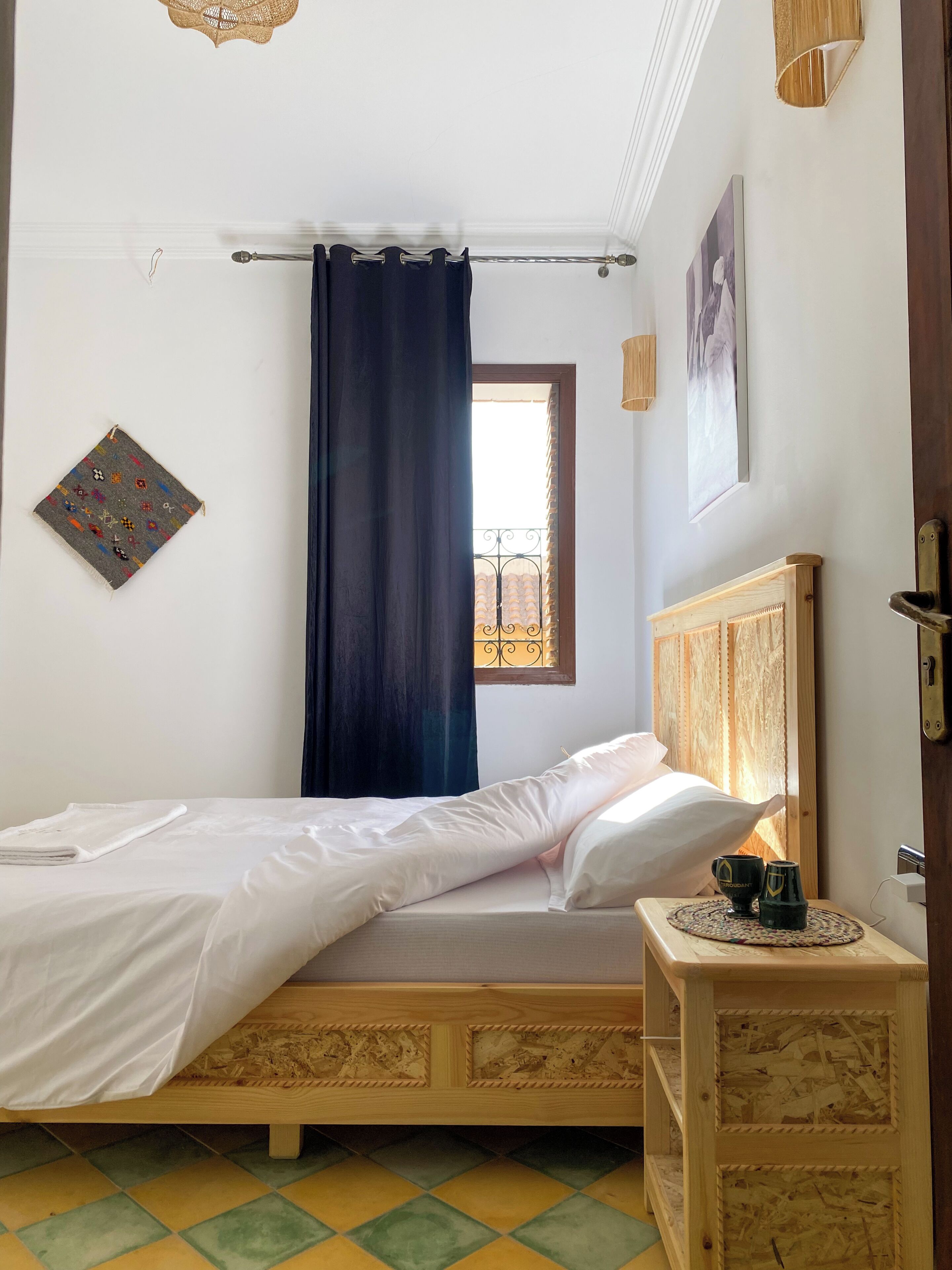 Un dormitorio con una cama, una mesita de noche con lámpara, una ventana con cortinas y un cuadro colgado en la pared.