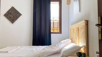 Un dormitorio con una cama, una mesita de noche con lámpara, una ventana con cortinas y un cuadro colgado en la pared.