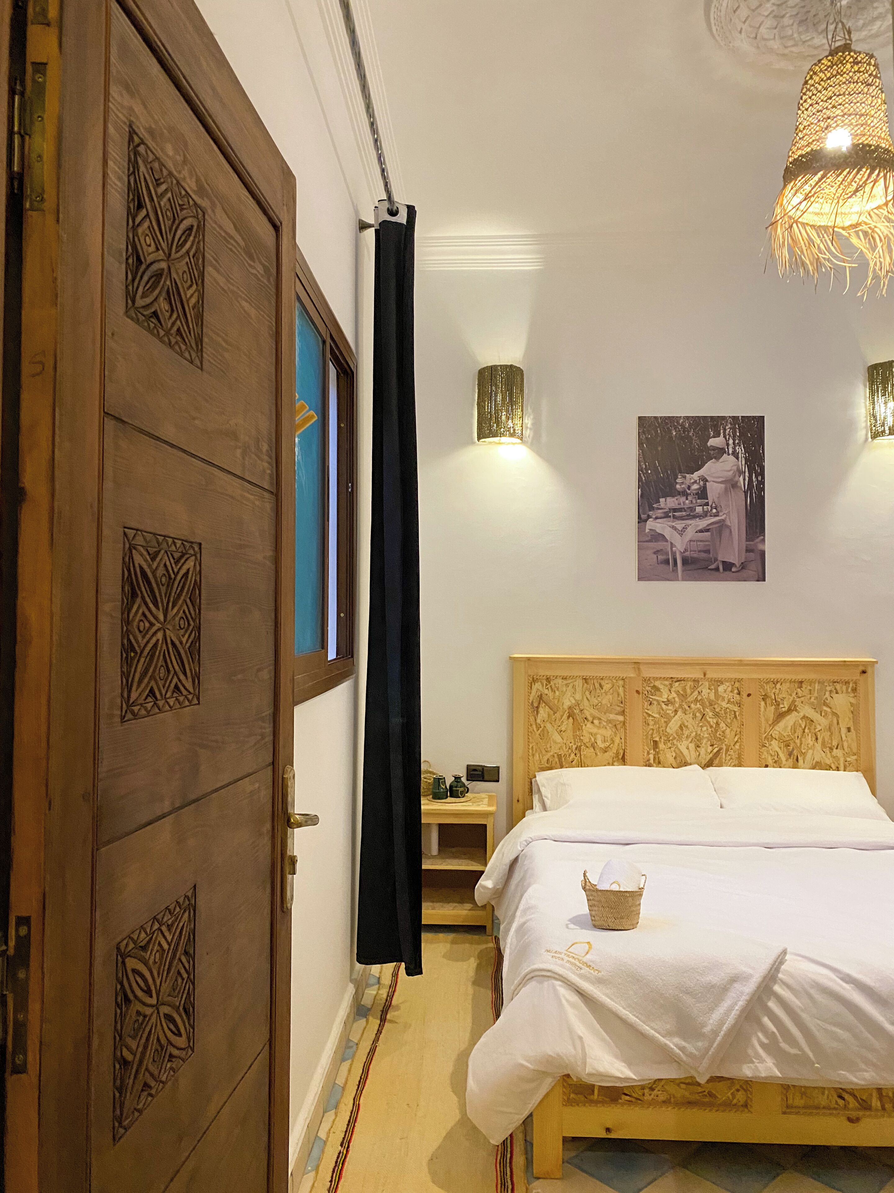 A bedroom with a wooden door, a bed with a white bedspread, a nightstand, a wall-mounted light, and a hanging light fixture.