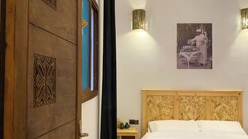 A bedroom with a wooden door, a bed with a white bedspread, a nightstand, a wall-mounted light, and a hanging light fixture.