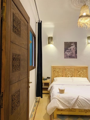 A bedroom with a wooden door, a bed with a white bedspread, a nightstand, a wall-mounted light, and a hanging light fixture. - Palais Taroudant (Taroudant)