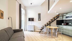 Un espace de vie moderne comprenant un canapé gris, une table à manger avec des chaises transparentes, un escalier, un coin cuisine et un téléviseur à écran plat fixé au mur.