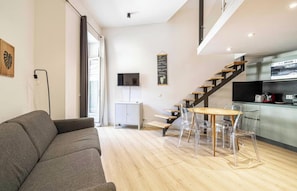 A modern living space with a grey sofa, a dining table with clear chairs, a staircase, a kitchen area, and a flat-screen TV mounted on the wall. - POSITIONED ON THE ICONIC PLACE GARIBALDI,IN THE MOST VIBRANT &HISTORIC SQUARES (Nice)