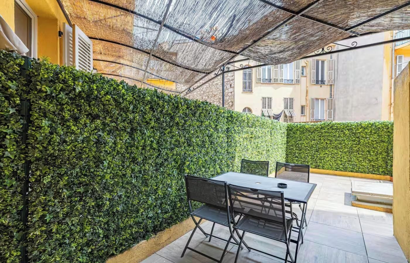Eine Terrasse im Freien mit Tisch und Stühlen, umgeben von einer Hecke und einer Struktur mit einem Reetdach.