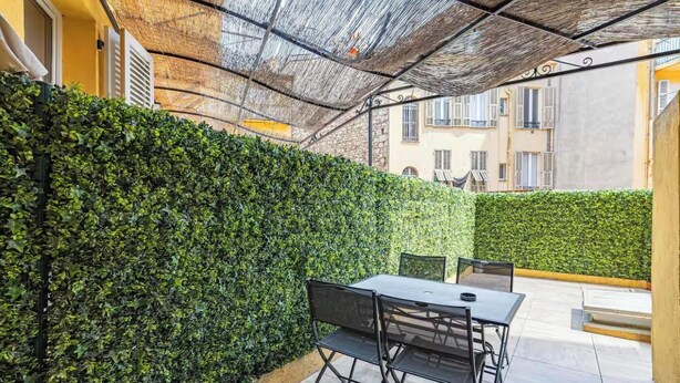 Une terrasse extérieure avec une table et des chaises, entourée d’une haie et d’une structure dotée d’un toit de chaume.