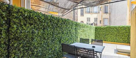 Un patio exterior con una mesa y sillas, rodeado por un seto y una estructura con techo de paja.