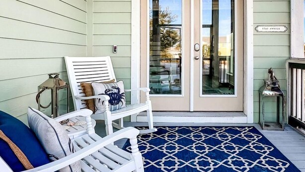 A porch with two white chairs, a blue patterned rug, and a door with a 'Stay Awhile' sign. - Arlanymor (Port St. Joe)