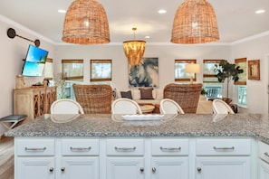 A kitchen with a granite countertop, white cabinets, wicker chairs, and a television on the wall. - Front and Sea (Port St. Joe)