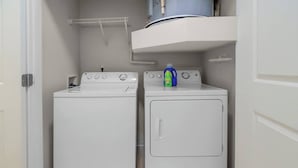 A laundry room with two white washing machines and a dryer, a metal shelf above the dryer, and a white door. - 2BR/2BA Luxury | Walkable + Parking | PET Friendly (Charlotte)