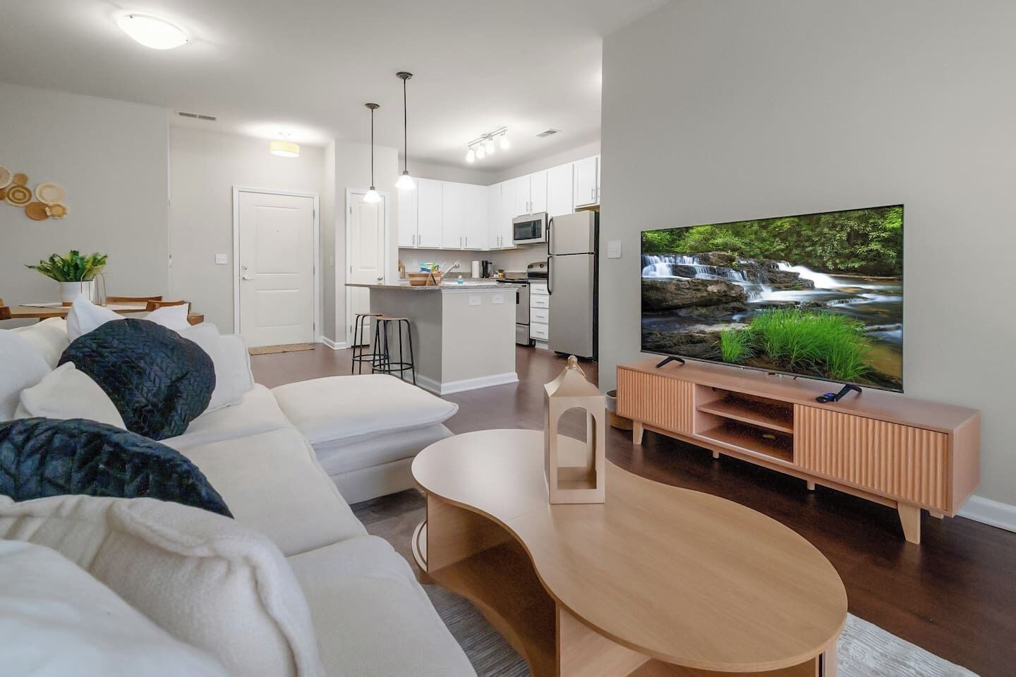 A modern living room with a sofa, a coffee table, a TV on a wooden stand, and a kitchen in the background.