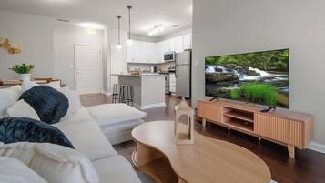 A modern living room with a sofa, a coffee table, a TV on a wooden stand, and a kitchen in the background.