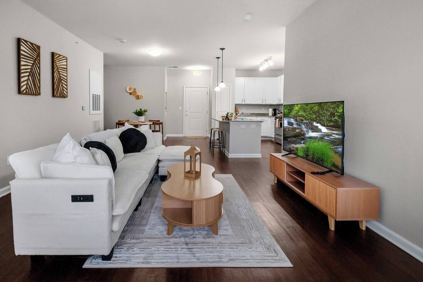 A modern living room with a white sofa, a coffee table, a TV on a wooden stand, and a kitchen in the background.