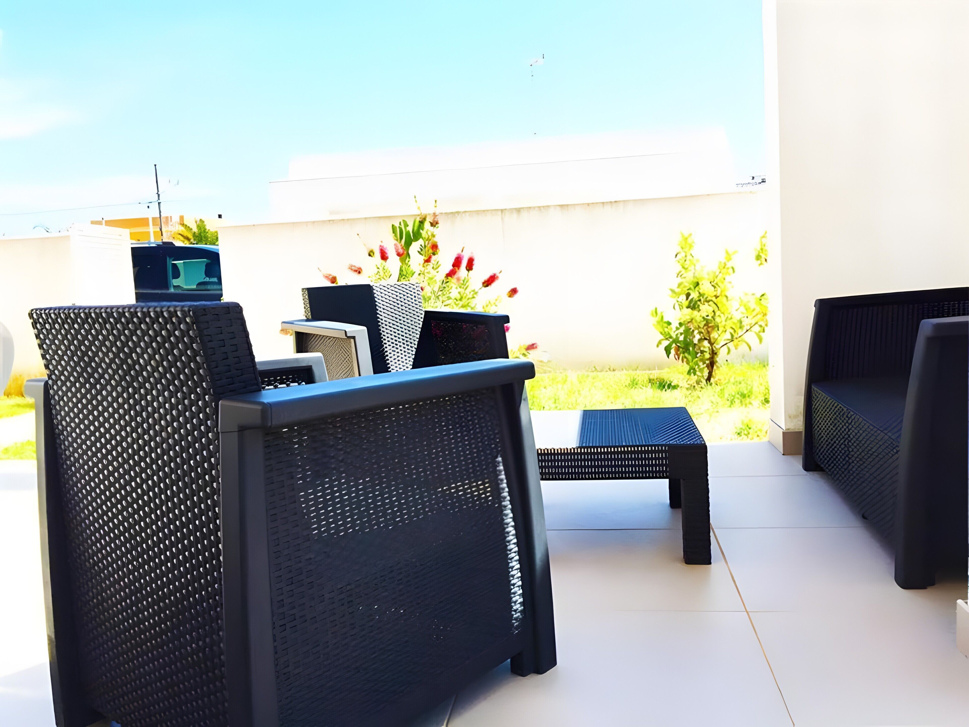 An outdoor seating area with wicker furniture and a small table, a flower arrangement, and a view of a building and greenery.