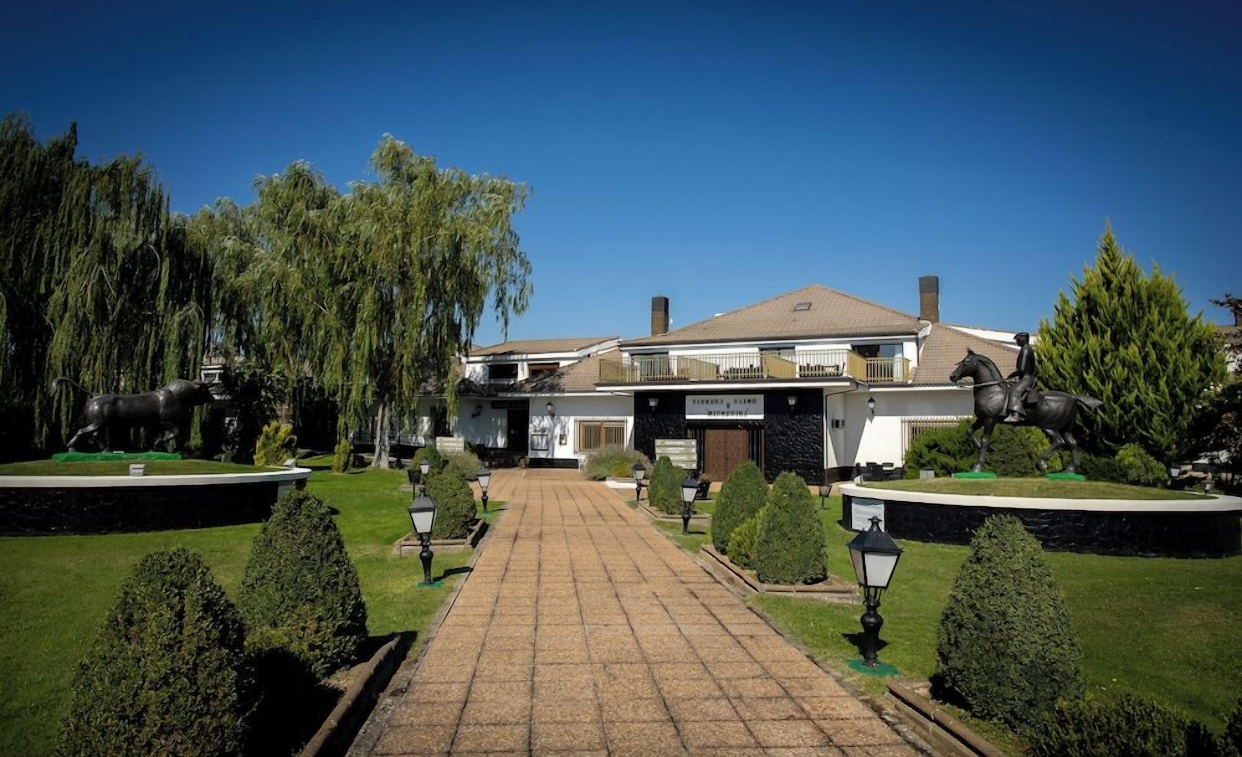 Un edificio señorial con una entrada central, rodeado de jardines cuidados y un camino empedrado que conduce a la entrada.