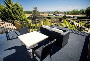 Un patio al aire libre con una mesa y sillas, vistas a un jardín ajardinado y una masa de agua distante.