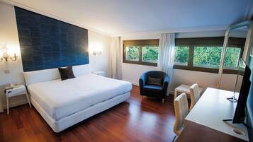 A modern hotel room with a large bed, a desk with a chair, a small table, and a window with curtains.