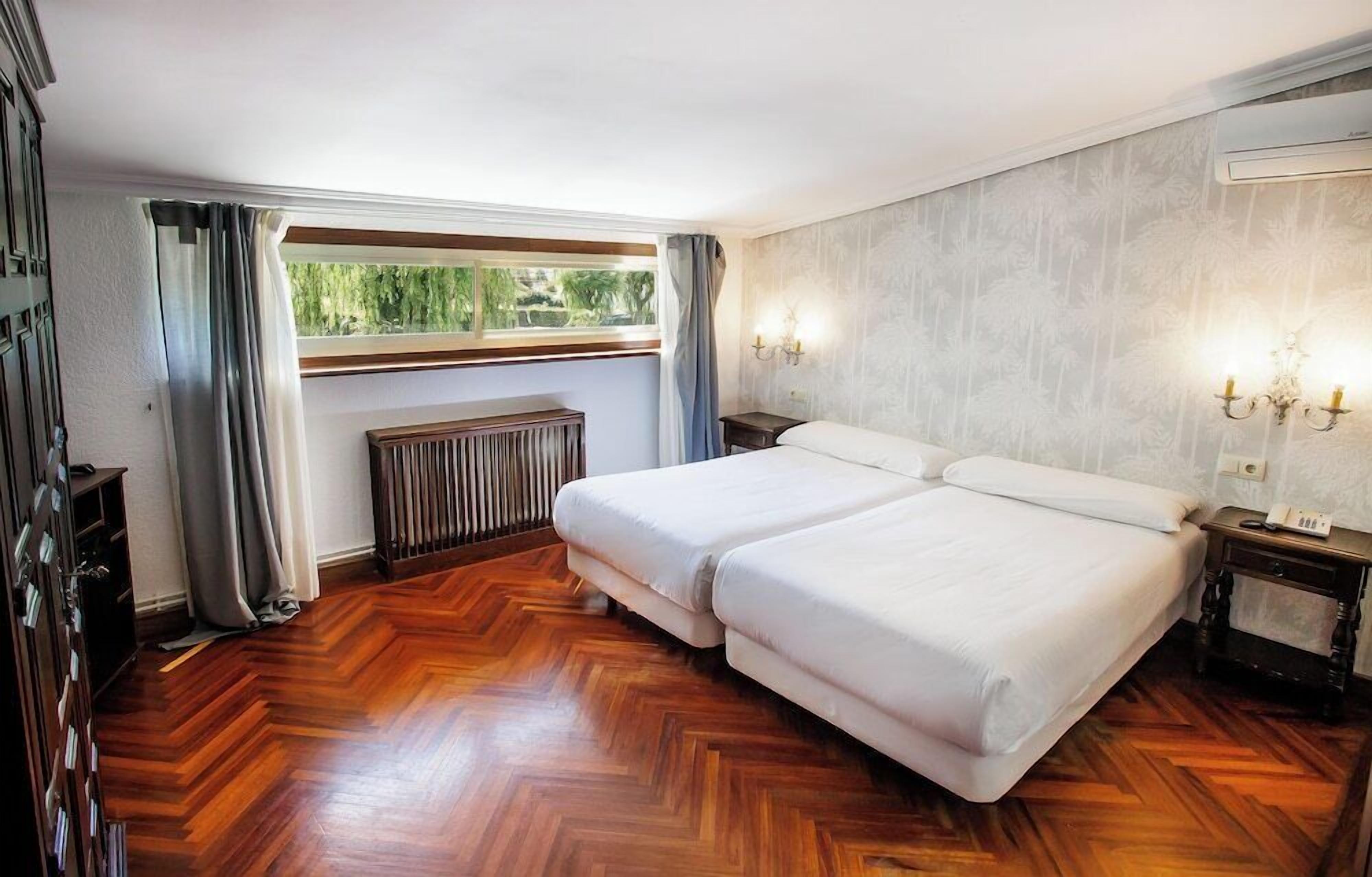 A hotel room with a double bed, a radiator, a window with curtains, and a wall-mounted light fixture.