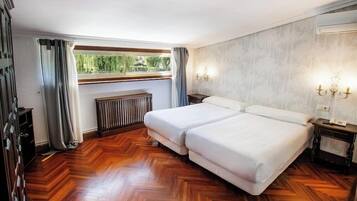 A hotel room with a double bed, a radiator, a window with curtains, and a wall-mounted light fixture.