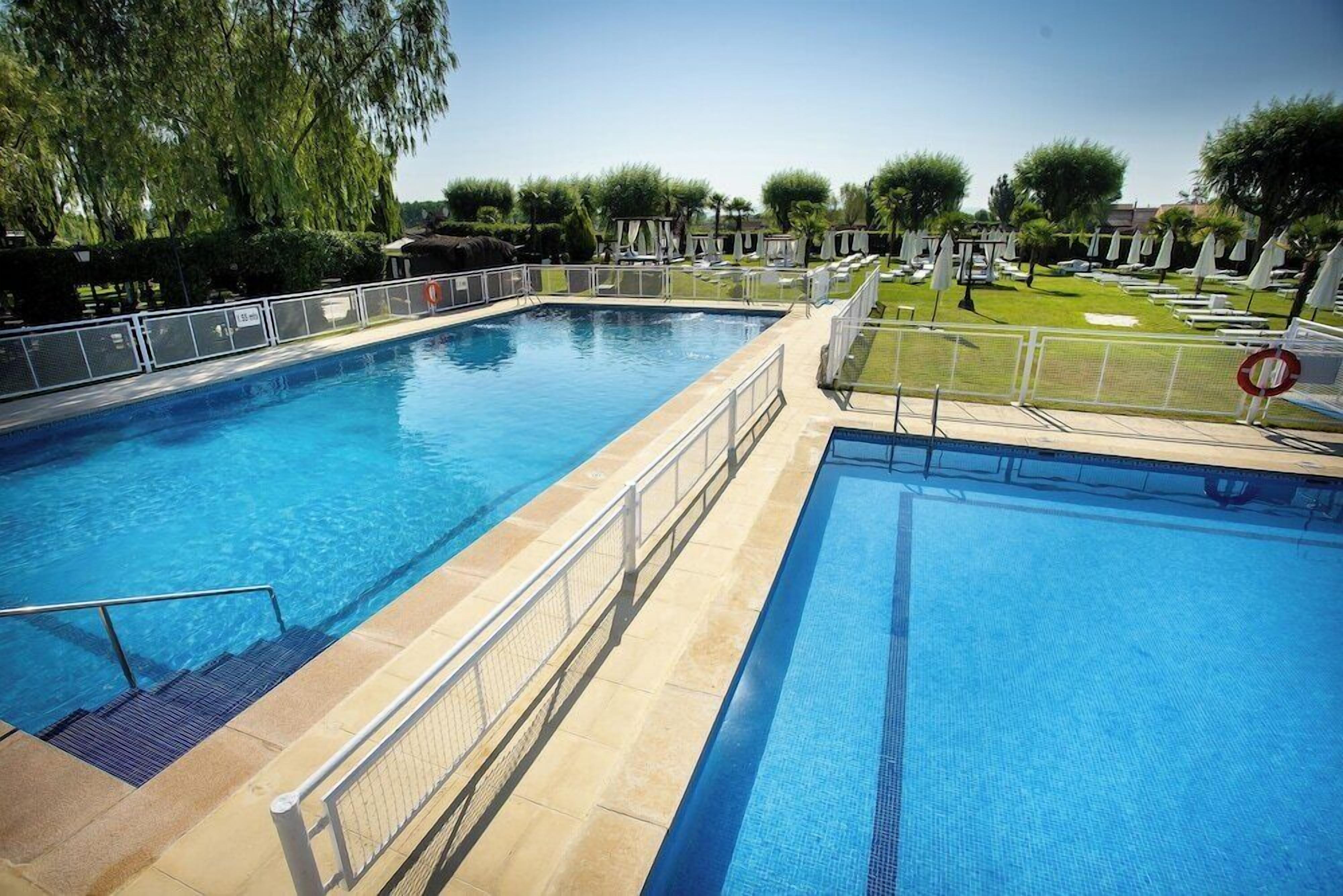 Una piscina con agua cristalina azul, rodeada por una cerca y un área de césped con sombrillas.