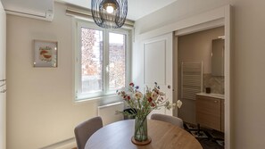 A dining area with a round table, chairs, a vase with flowers, a window with a view of a brick building, and a wall-mounted air conditioner. - Perchoir d'Augustin Arras • calme • CLIM • centre (Arras)