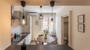 A modern kitchen with pendant lighting, a dining area with a round table, and a view of a brick building outside. - Perchoir d'Augustin Arras • calme • CLIM • centre (Arras)