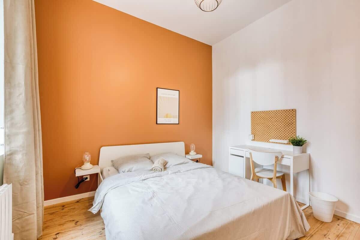 A bedroom with a bed, bedside table, desk, chair, and a small trash bin.