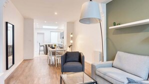Un salon moderne avec un canapé, un fauteuil et une table basse, un coin repas avec des chaises, et une cuisine en arrière-plan.