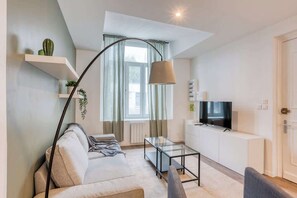 A modern living room with a sofa, a glass coffee table, a TV on a white cabinet, and a floor lamp.