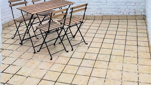 Une terrasse avec un mur en briques blanches, un sol carrelé et une table en bois entourée de chaises en métal.