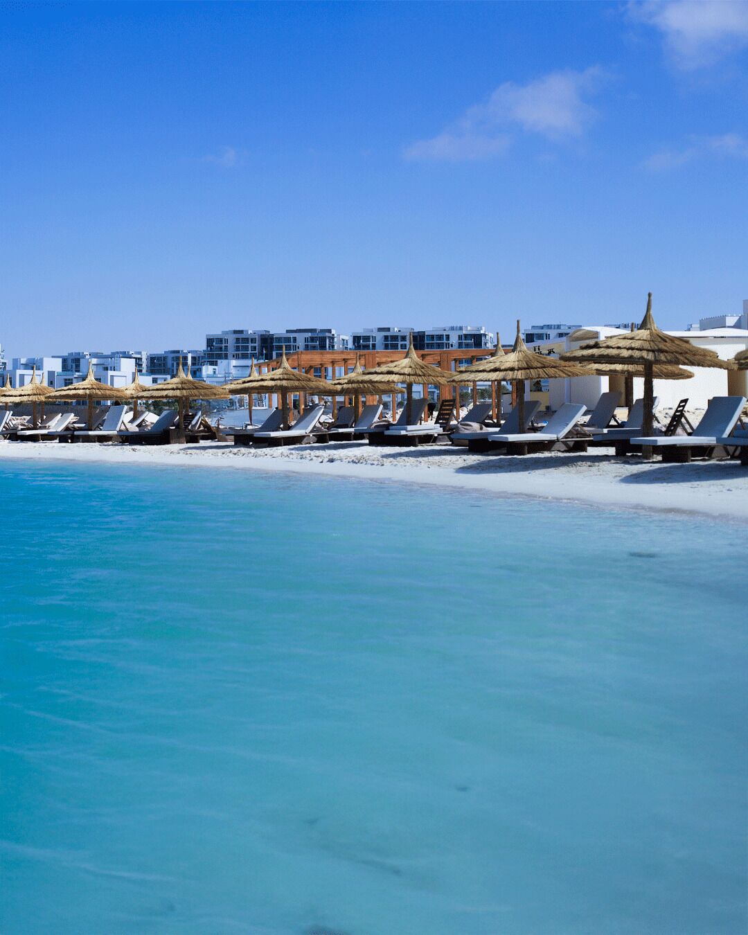 Un hotel frente a la playa con tumbonas y sombrillas de paja, edificios blancos al fondo.