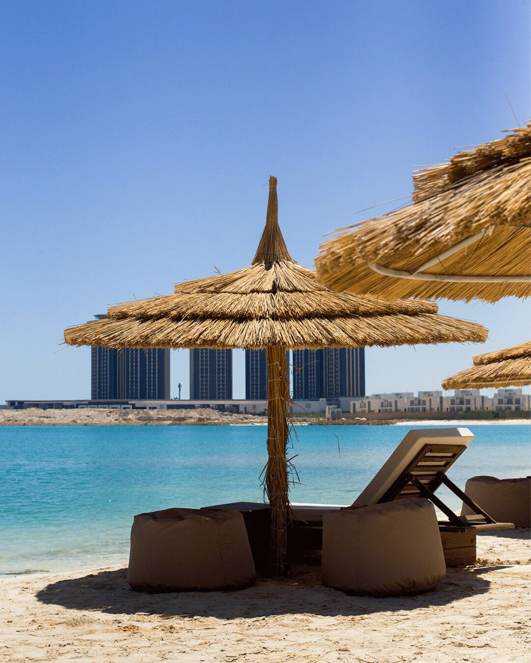 A beach with thatched umbrellas, lounge chairs, and a view of high-rise buildings in the background.