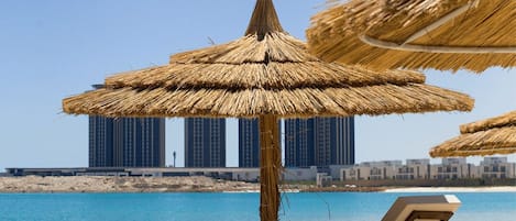 En strand med parasoller av halm, solstolar och utsikt över höga byggnader i bakgrunden.